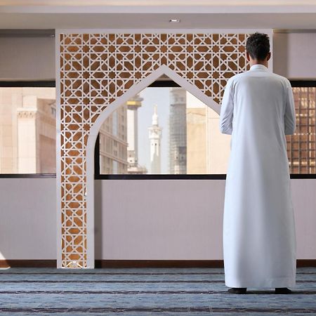 Makarem Ajyad Makkah Hotel Mekka Zewnętrze zdjęcie The photo depicts a person standing with their back facing the viewer, wearing a traditional white garment. They are in a spacious interior setting that features an ornate archway with intricate patterns, typical of Islamic architecture. Through the 
