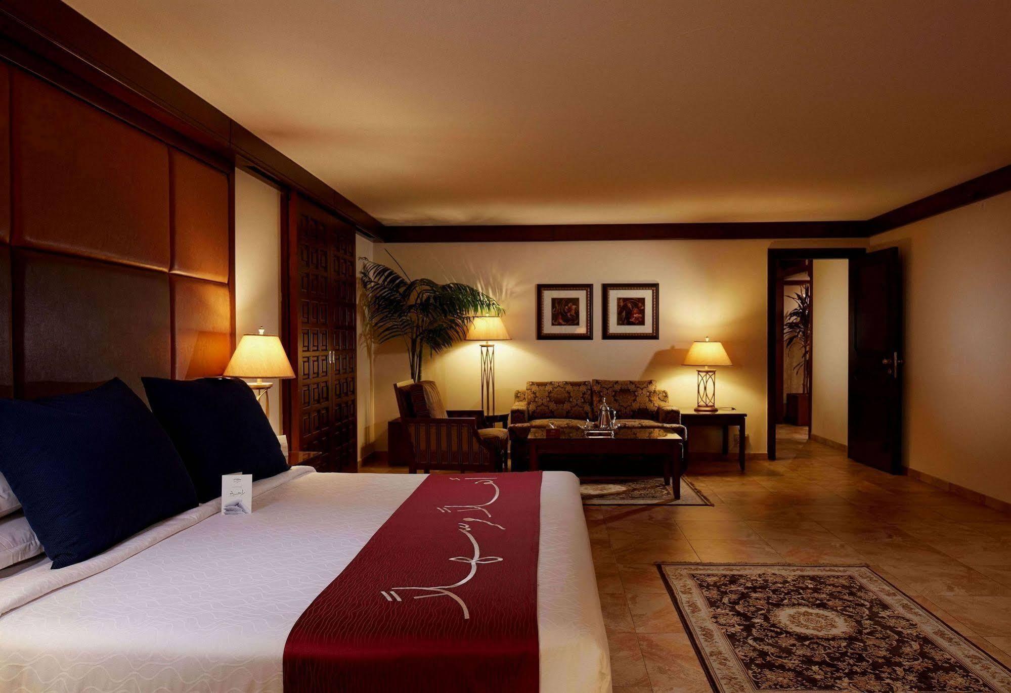Makarem Ajyad Makkah Hotel Mekka Zewnętrze zdjęcie The photo depicts a cozy hotel room with a warm and inviting atmosphere. It features a large bed with dark blue pillows and a maroon throw blanket. On either side of the bed, there are lamps providing soft lighting. In the background, a small seating