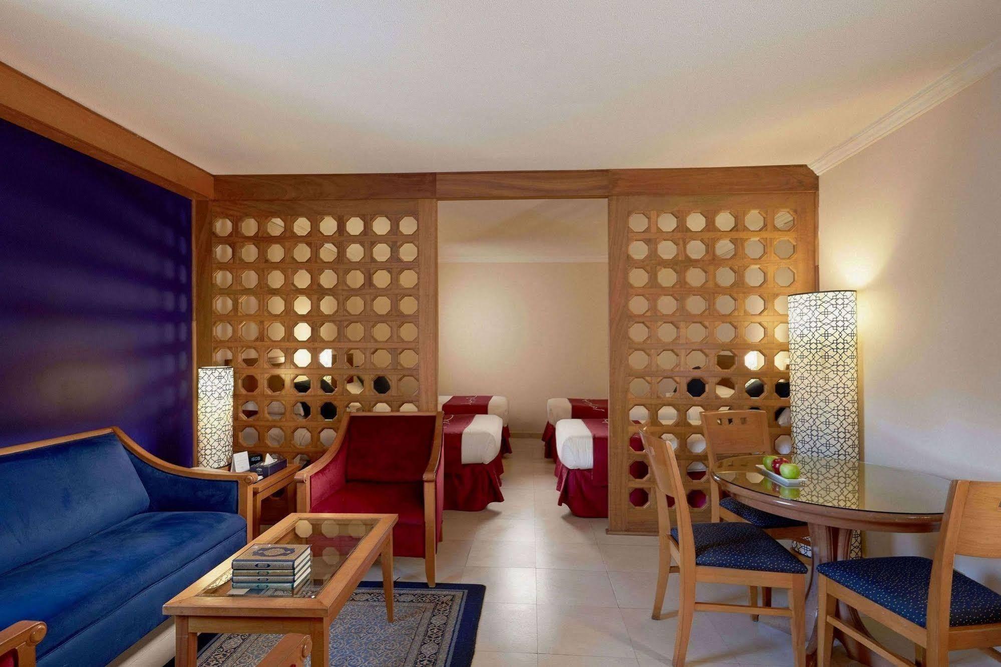 Makarem Ajyad Makkah Hotel Mekka Zewnętrze zdjęcie The photo shows a well-furnished hotel room or suite. On the left side, there is a seating area featuring a blue sofa and a small coffee table, as well as a couple of decorative items. The room is separated by a decorative wooden partition with circu