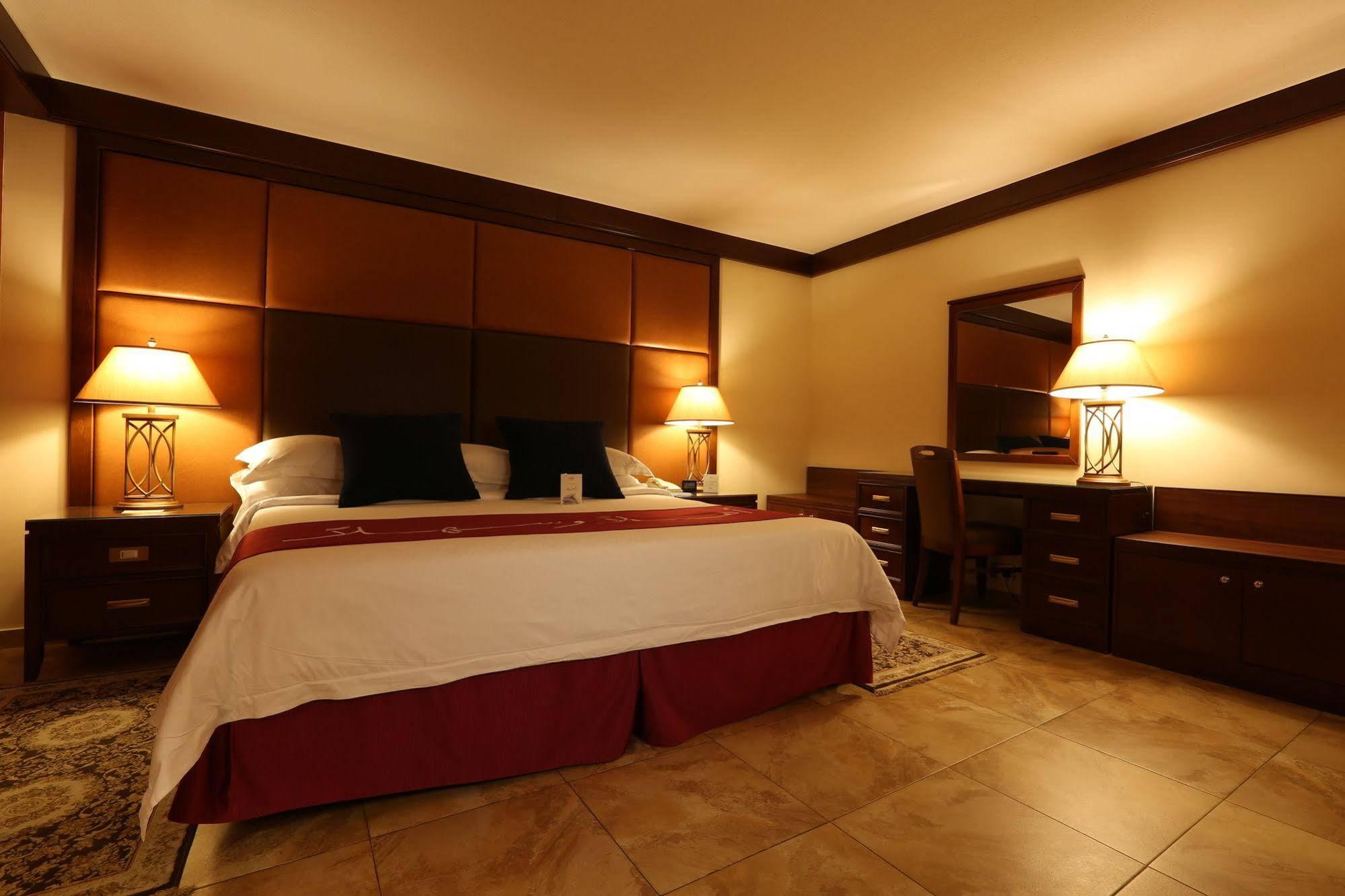 Makarem Ajyad Makkah Hotel Mekka Zewnętrze zdjęcie The photo shows a well-appointed hotel room with a cozy and inviting atmosphere. It features a large bed with a white comforter and dark pillows, positioned against a wooden headboard. There are two bedside lamps providing soft lighting on either sid