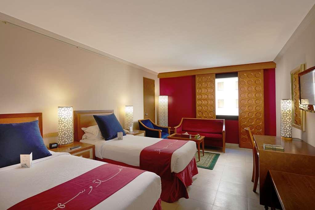 Makarem Ajyad Makkah Hotel Mekka Pokój zdjęcie The photo shows a hotel room featuring two twin beds with red and white bedding. There are decorative lamps on either side of the beds. A small seating area is visible, including a blue chair and a red sofa. There is a wooden desk against one wall, a