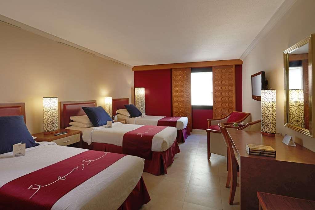 Makarem Ajyad Makkah Hotel Mekka Pokój zdjęcie The photo shows a hotel room featuring three beds arranged with red and white bedding. The walls are painted in a neutral tone, and there are decorative lamps on nightstands beside each bed, providing ambient lighting. One of the walls has a window c