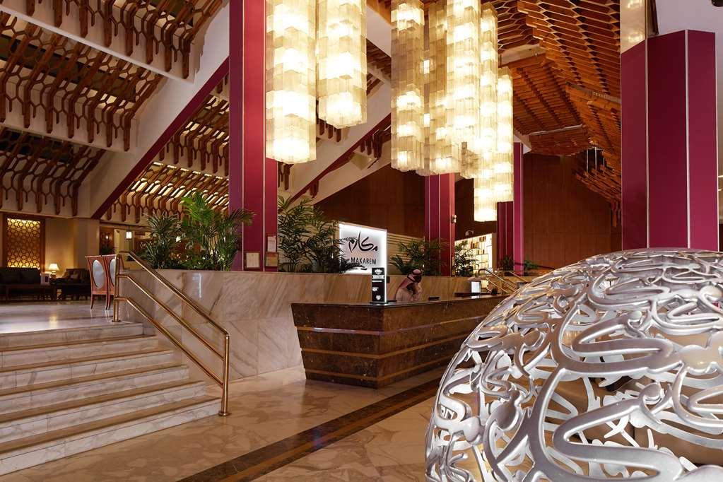 Makarem Ajyad Makkah Hotel Mekka Wnętrze zdjęcie The photo depicts a luxurious hotel lobby. It features a grand entrance with a staircase leading up to a reception area. The lobby has high ceilings adorned with striking, modern light fixtures that hang down, providing a warm ambiance. The walls are