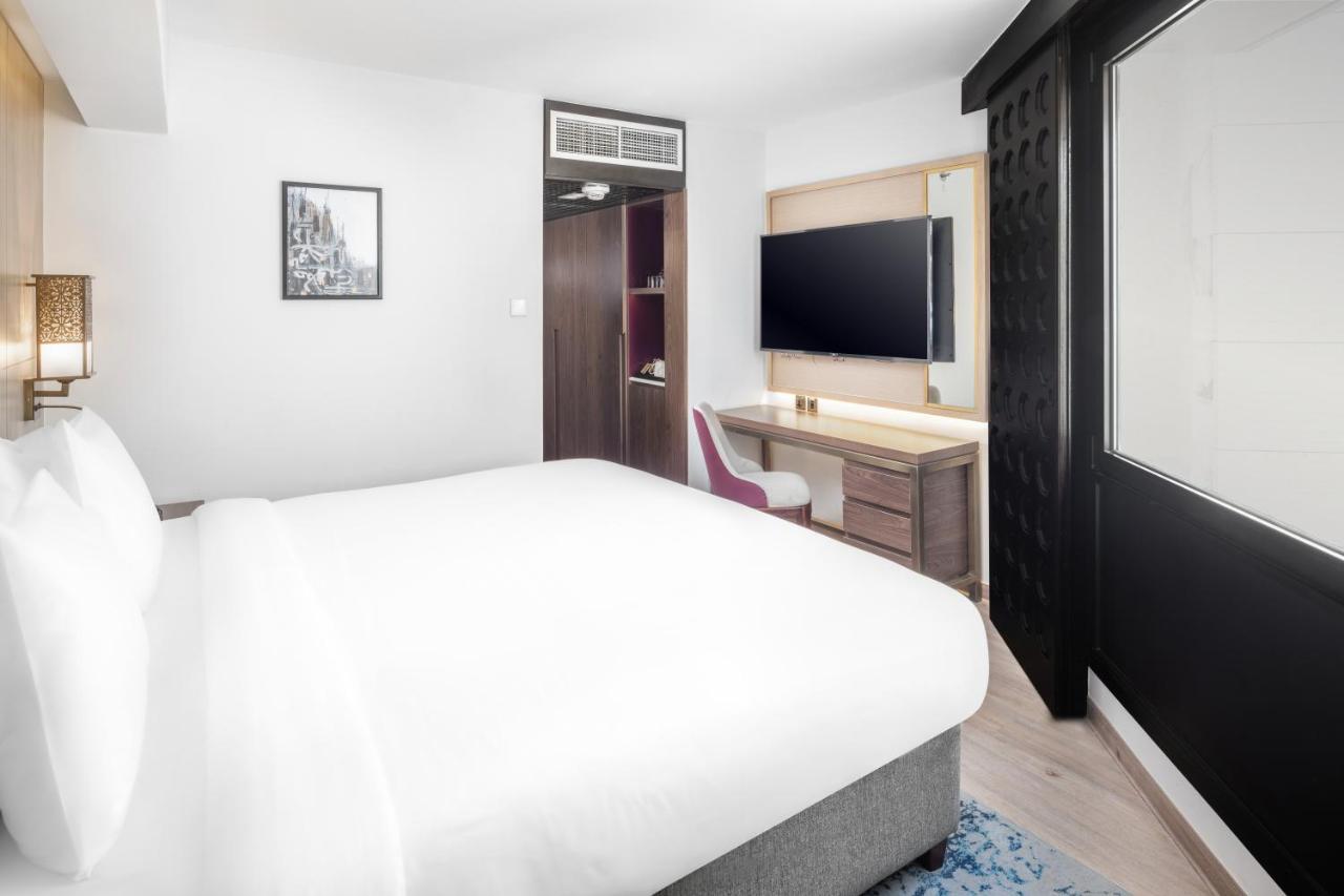Makarem Ajyad Makkah Hotel Mekka Zewnętrze zdjęcie The photo shows a modern hotel room featuring a large bed with white linens at the forefront. In the background, there is a wall-mounted television across from a desk with a chair. A wardrobe or cabinet is located to the side, and there's a decorativ