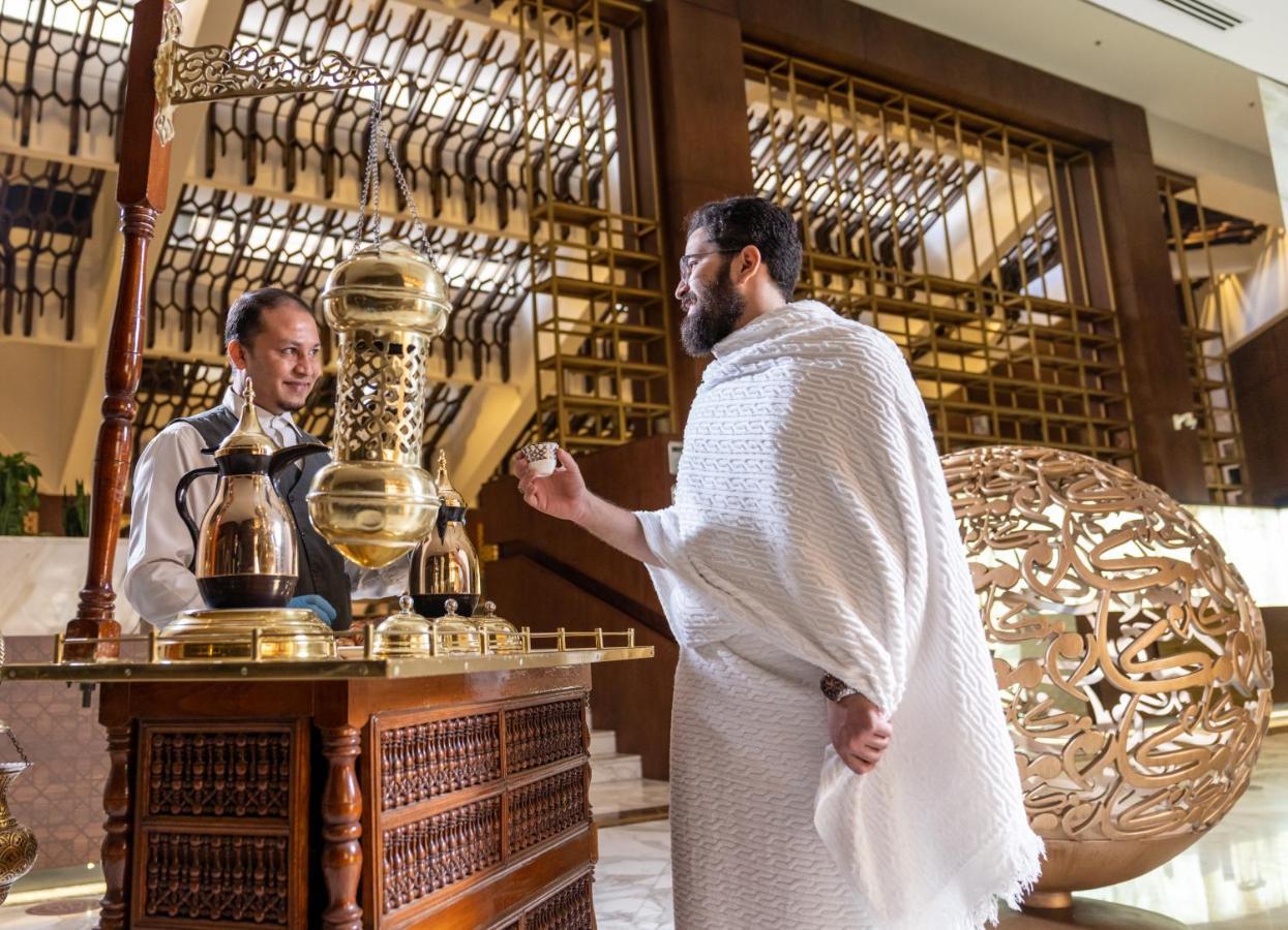 Makarem Ajyad Makkah Hotel Mekka Zewnętrze zdjęcie The photo depicts a scene in a luxurious setting, likely a hotel or a fine dining establishment. In the foreground, a man wearing traditional attire, possibly a white garment, is engaging with a staff member. The staff member is behind a wooden count