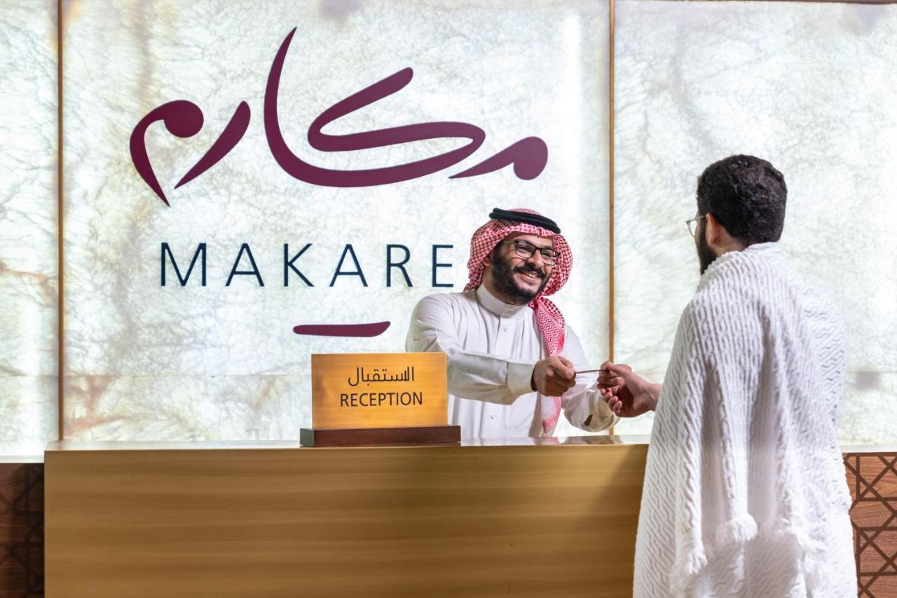 Makarem Ajyad Makkah Hotel Mekka Zewnętrze zdjęcie The photo shows a reception area with a sign that reads "MAKARE" in both Arabic and English. In the foreground, a staff member wearing traditional attire is smiling and shaking hands with a visitor, who is clad in a white garment, typical for formal 