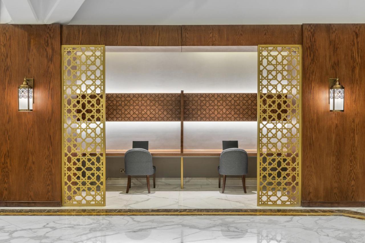 Makarem Ajyad Makkah Hotel Mekka Zewnętrze zdjęcie The photo shows an elegantly designed space that features a reception or working area. There are two workstations with chairs positioned in front of them. The backdrop has a decorative panel with a geometric pattern. The area is framed by ornate gold