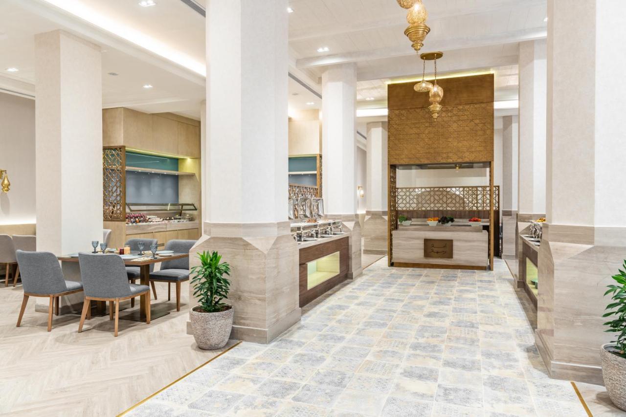 Makarem Ajyad Makkah Hotel Mekka Zewnętrze zdjęcie The photo depicts a modern, elegant dining area, likely inside a restaurant or hotel. It features a spacious layout with tall ceilings and large columns. The flooring is a light-colored tiled design. 

To one side, there are several dining tables and