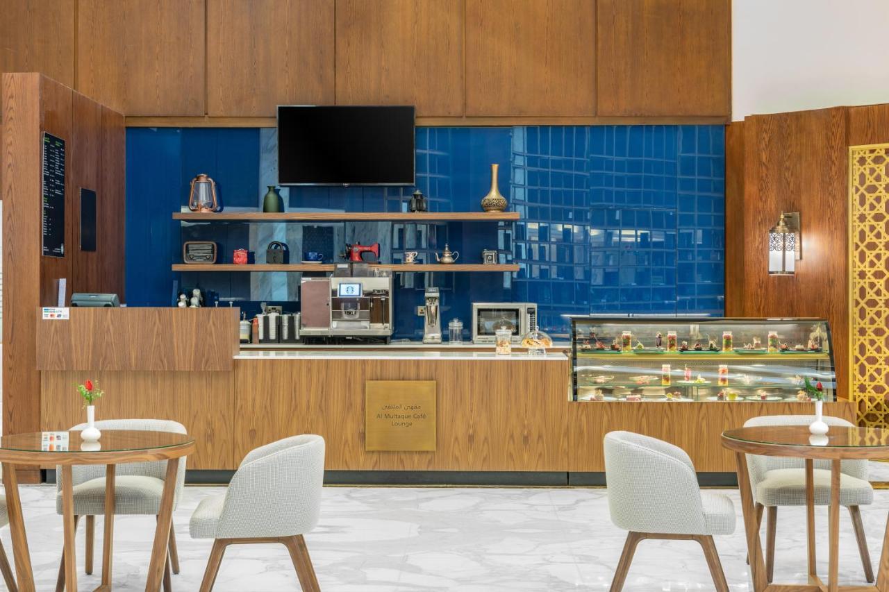 Makarem Ajyad Makkah Hotel Mekka Zewnętrze zdjęcie The photo shows the interior of a café. The design features warm wood paneling and a striking blue tiled wall. In the foreground, there are several light-colored chairs and small tables arranged neatly. Behind the counter, there's a variety of coffee