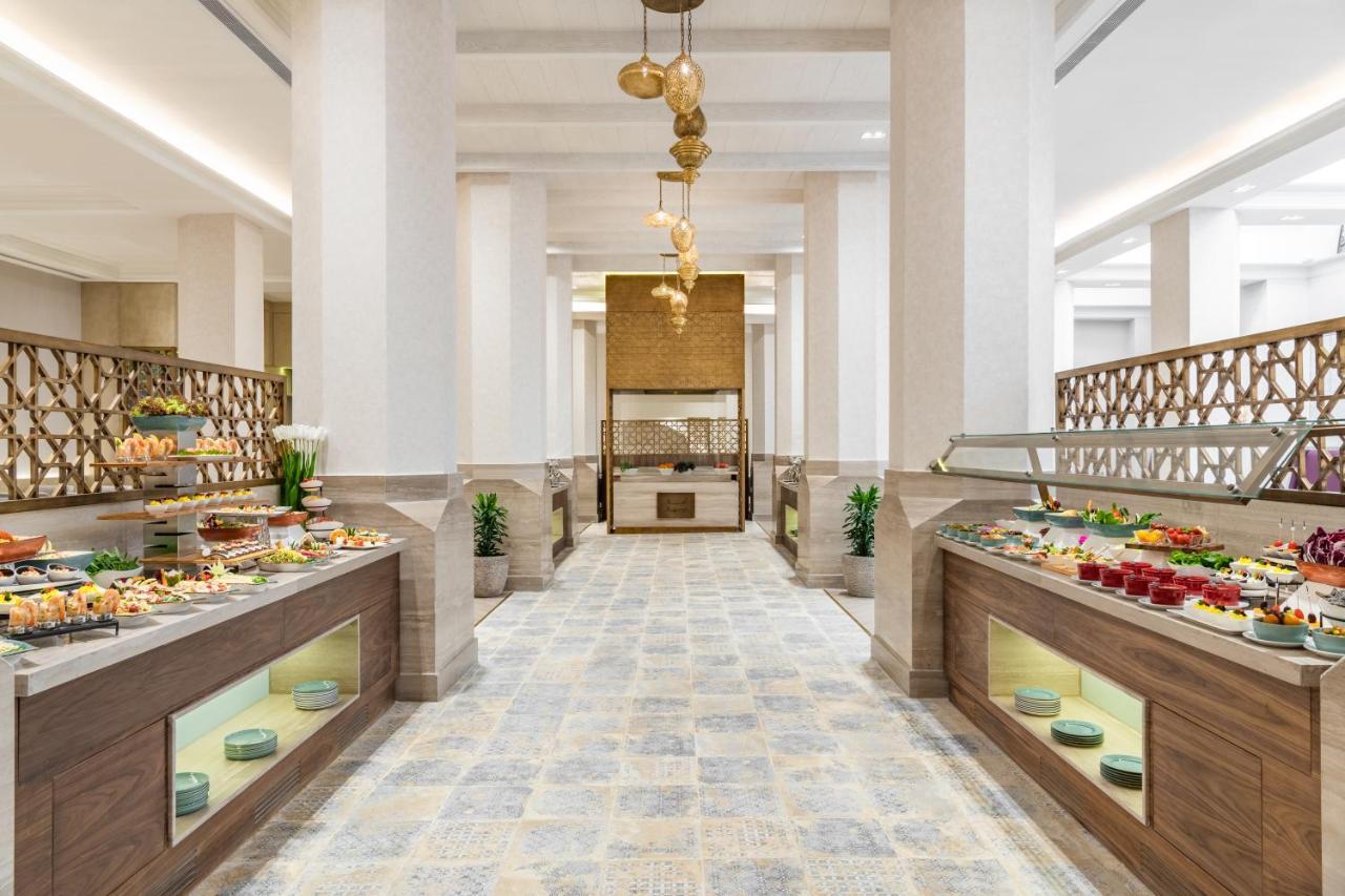 Makarem Ajyad Makkah Hotel Mekka Zewnętrze zdjęcie The photo shows an elegant restaurant or buffet area. There are two long buffet tables on either side, displaying a variety of colorful dishes and desserts. The tables have an organized layout and are adorned with small plants. In the center of the r