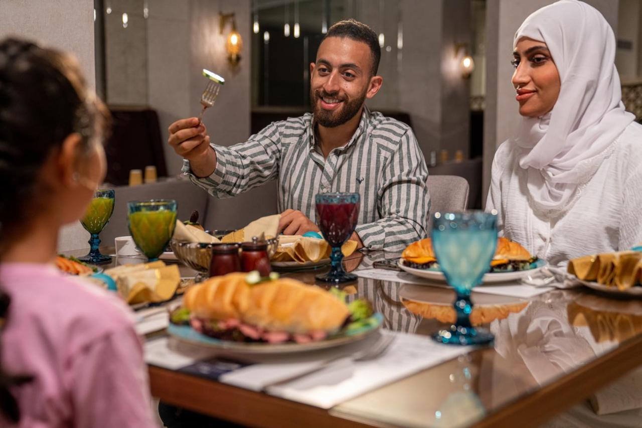 Makarem Ajyad Makkah Hotel Mekka Zewnętrze zdjęcie The photo shows a dining scene with three people enjoying a meal together. A man is seated at the table, smiling and holding a utensil, possibly ready to serve or share food. He is dressed in a striped shirt. Across from him is a woman wearing a whit