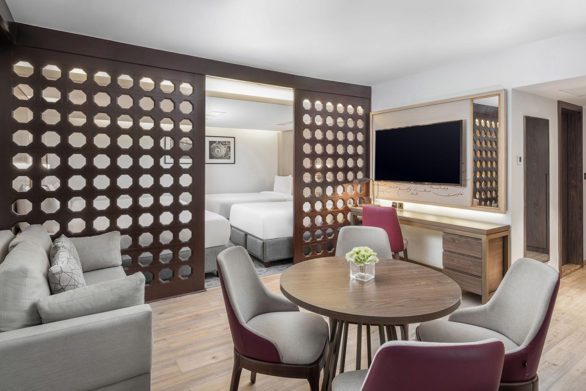 Makarem Ajyad Makkah Hotel Mekka Zewnętrze zdjęcie The photo shows a modern hotel room or suite. It features a living area with a comfortable gray sofa and a round wooden dining table surrounded by upholstered chairs. A large flat-screen TV is mounted on the wall opposite the seating area. There's a 