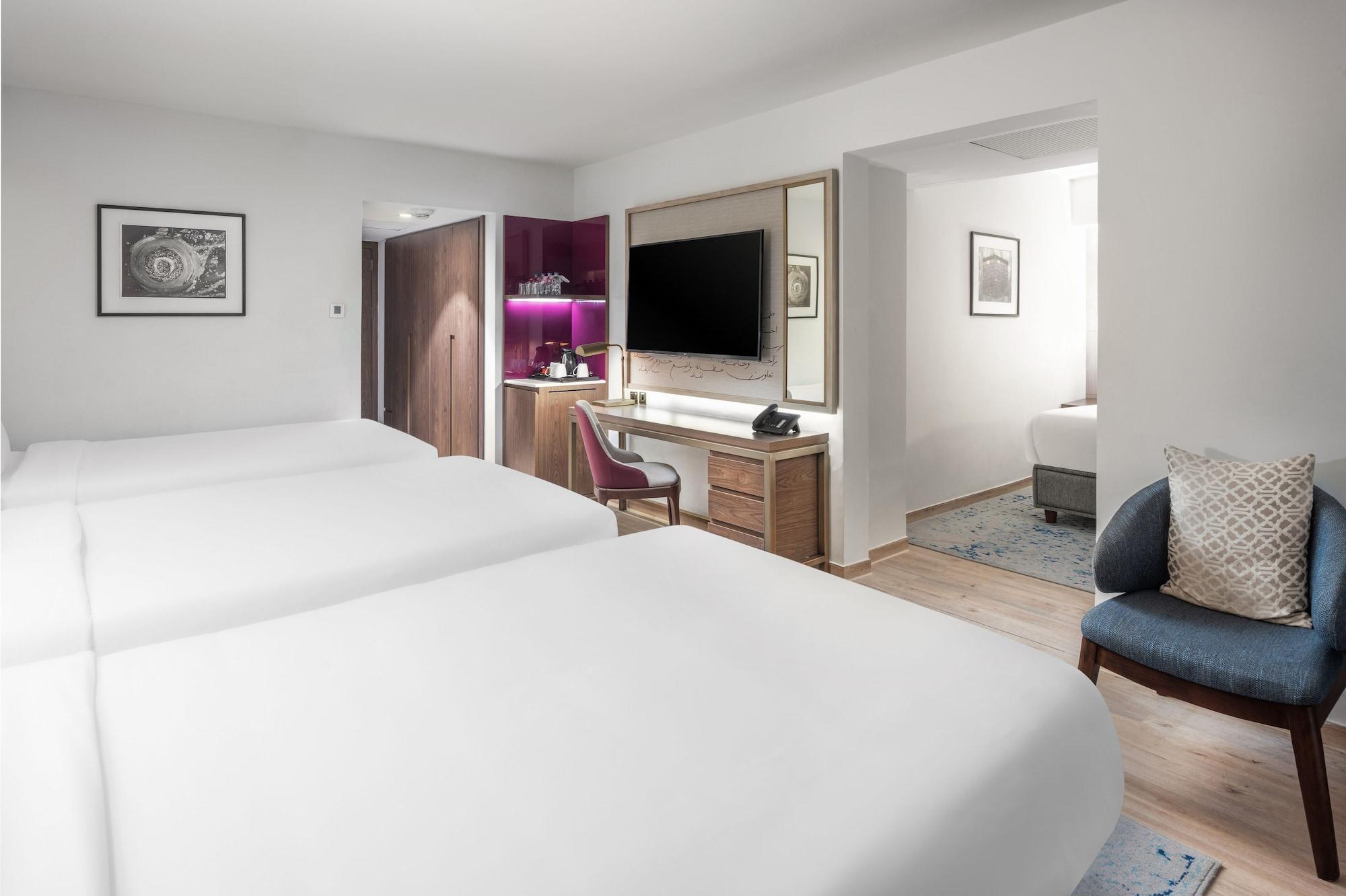 Makarem Ajyad Makkah Hotel Mekka Zewnętrze zdjęcie The photo shows a modern hotel room featuring two white beds with smooth linens. The room has a minimalistic design with light-colored walls and wooden flooring. On one side, there is a desk and chair next to a television mounted on the wall, along w