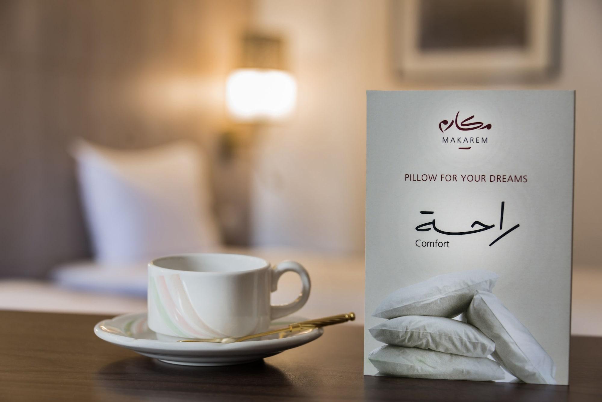 Makarem Ajyad Makkah Hotel Mekka Zewnętrze zdjęcie The image features a cozy setting, likely a hotel room. In the foreground, there is a white cup of coffee or tea placed on a small saucer, accompanied by a gold spoon. Beside the cup is a decorative card or sign that reads "Pillow for Your Dreams" an