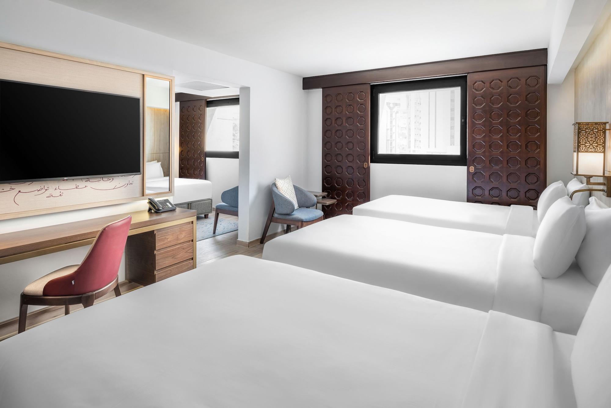 Makarem Ajyad Makkah Hotel Mekka Zewnętrze zdjęcie The photo shows a hotel room featuring three beds with white linens. There is a large flat-screen TV mounted on the wall opposite the beds, and a wooden side table beneath it. A red chair is positioned near the TV, while a blue upholstered chair is p
