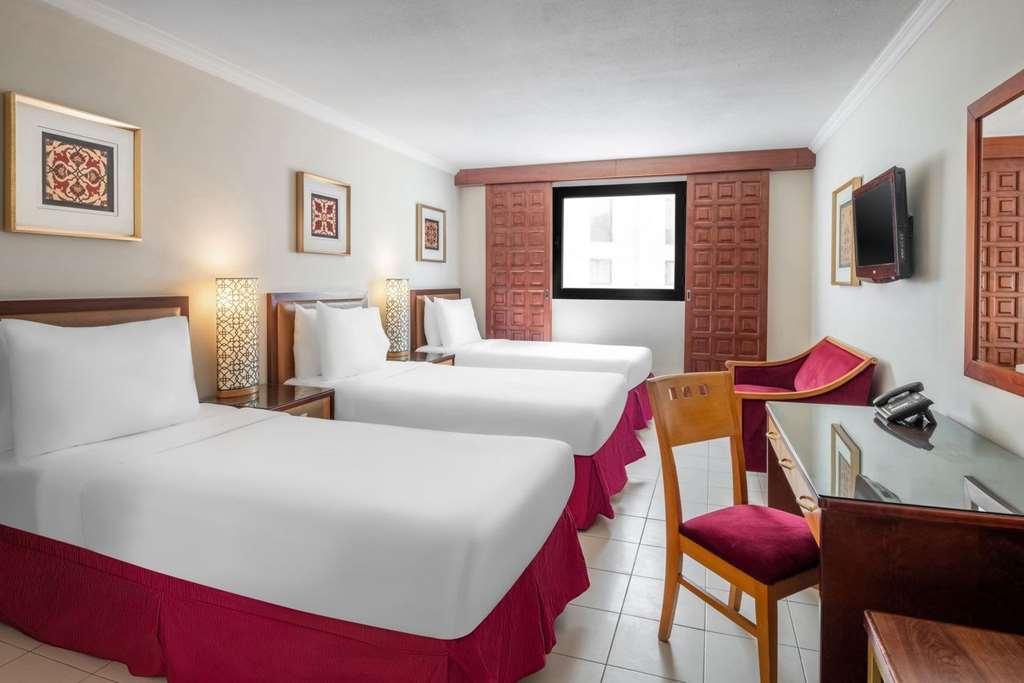 Makarem Ajyad Makkah Hotel Mekka Pokój zdjęcie The photo shows a hotel room featuring two twin beds with white linens and red bed skirts. There are decorative elements on the walls, including framed artwork. A large window provides natural light, while a desk and a chair are positioned next to it