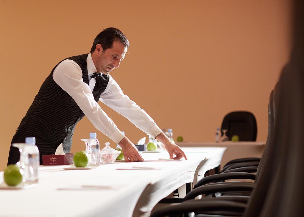 Makarem Ajyad Makkah Hotel Mekka Zewnętrze zdjęcie The image shows a waiter or service staff member in a formal attire, which includes a vest and bow tie. He is preparing a long table, likely for an event or meeting. The table is set with items such as water bottles and green apples, and there are se